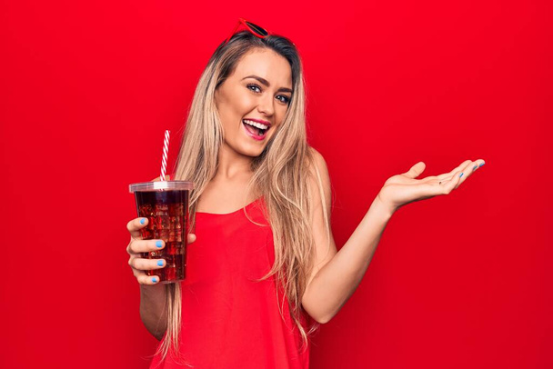 Krásná blondýnka pití cola nápoj občerstvení pomocí slámy nad červeným pozadím slaví úspěch s veselým úsměvem a vítězný výraz se zvednutou rukou - Fotografie, Obrázek