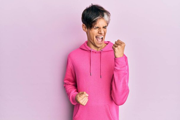 Νεαρός Ισπανόφωνος με ροζ φούτερ γιορτάζει έκπληκτος και έκπληκτος για την επιτυχία με τα χέρια ψηλά και τα μάτια κλειστά  - Φωτογραφία, εικόνα