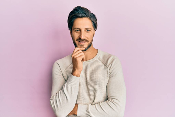Young hispanic man wearing casual winter sweater looking confident at the camera with smile with crossed arms and hand raised on chin. thinking positive.  - Photo, Image