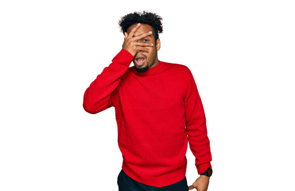 Fiatal afro-amerikai férfi szakállal alkalmi téli pulóvert visel leselkedik sokk borító arc és a szemek kézzel, néz keresztül ujjak zavarba ejtő kifejezés.  - Fotó, kép