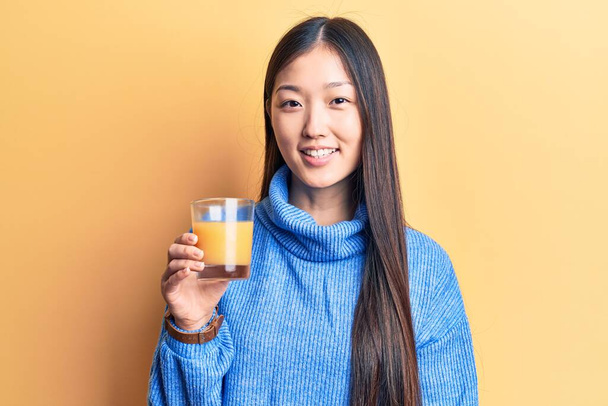 Jovem mulher chinesa bonita bebendo copo de suco de laranja olhando positivo e feliz de pé e sorrindo com um sorriso confiante mostrando dentes  - Foto, Imagem