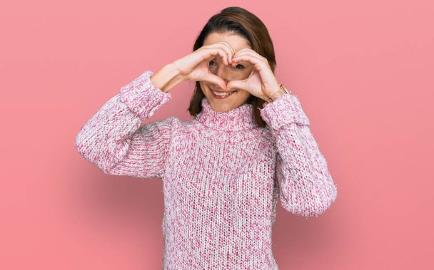 Joven chica caucásica con suéter de lana de invierno haciendo forma de corazón con la mano y los dedos sonriendo mirando a través de signo  - Foto, imagen