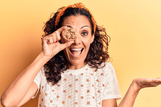 Mujer hermosa de mediana edad sosteniendo una galleta de chocolate sobre el ojo celebrando el logro con sonrisa feliz y expresión ganadora con la mano levantada  - Foto, imagen