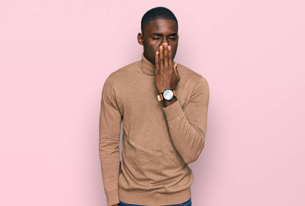 Fiatal afro-amerikai férfi alkalmi téli pulóvert visel unatkozva ásít fáradt eltakarja a száját kézzel. nyugtalanság és álmosság.  - Fotó, kép