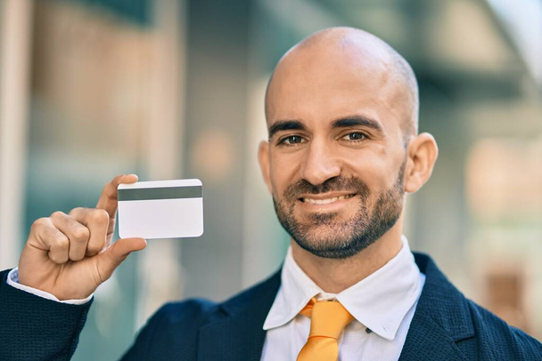 Joven empresario calvo hispano sonriendo feliz sosteniendo tarjeta de crédito en la ciudad. - Foto, imagen