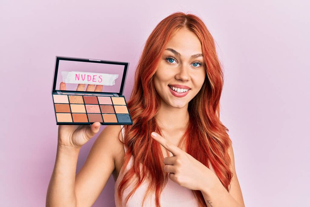 Young redhead woman holding makeup nudes smiling happy pointing with hand and finger  - Photo, Image