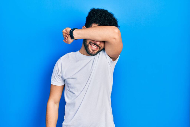 Young arab man with beard wearing casual white t shirt covering eyes with arm smiling cheerful and funny. blind concept.  - Foto, immagini