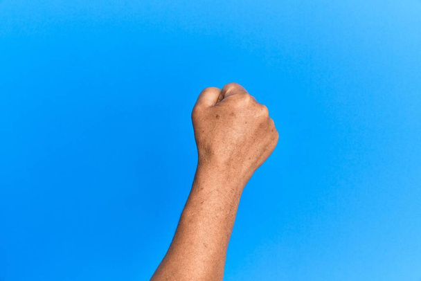 Χέρι του πρεσβύτερου ισπανόφωνου πάνω από το μπλε απομονωμένο υπόβαθρο κάνει διαμαρτυρία και χειρονομία επανάστασης, γροθιά εκφράζοντας δύναμη και δύναμη  - Φωτογραφία, εικόνα