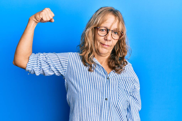 светловолосая женщина среднего возраста в повседневной одежде и очках сильного человека, демонстрирующего мышцы рук, уверенная и гордящаяся властью  - Фото, изображение