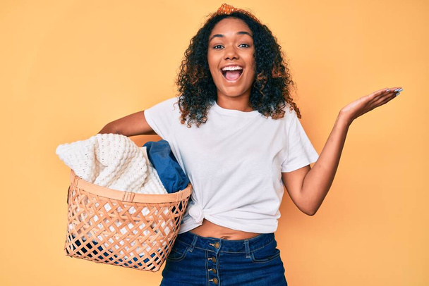Joven mujer afroamericana sosteniendo cesta de lavandería celebrando la victoria con sonrisa feliz y la expresión ganadora con las manos levantadas  - Foto, Imagen