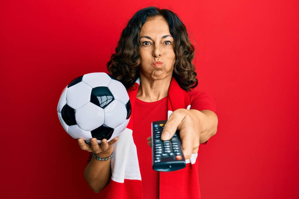 Латиноамериканка средних лет, футбольный хулиган, держит мяч, используя ТВ-контроль, пыхтя щеками со смешным лицом. рот надутый воздухом, ловящий воздух.  - Фото, изображение