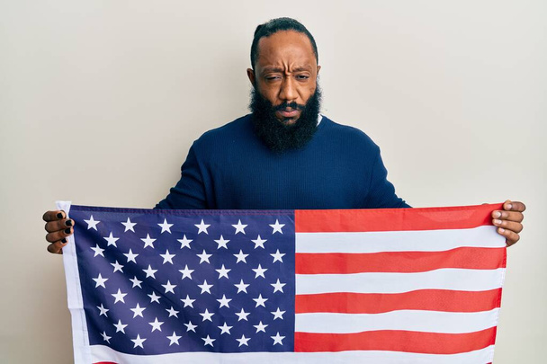 Junger afrikanisch-amerikanischer Mann, der die Flagge der Vereinigten Staaten in der Hand hält, ist skeptisch und nervös und runzelt ob des Problems die Stirn. negative person.  - Foto, Bild