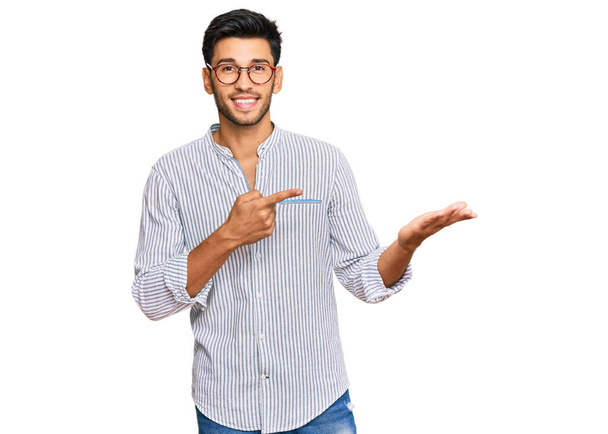 Νεαρός όμορφος άνδρας φορώντας καθημερινά ρούχα και γυαλιά έκπληκτος και χαμογελώντας στην κάμερα ενώ παρουσιάζει με το χέρι και δείχνοντας με το δάχτυλο.  - Φωτογραφία, εικόνα