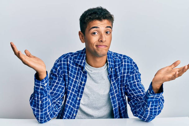 Young handsome african american man wearing casual clothes sitting on the table clueless and confused expression with arms and hands raised. doubt concept.  - Photo, Image