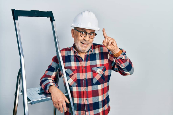 Handsome mature handyman close to construction stairs wearing hardhat smiling amazed and surprised and pointing up with fingers and raised arms.  - Fotografie, Obrázek