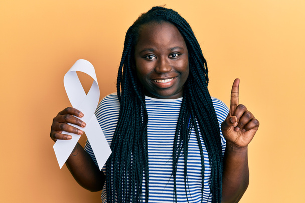 Young black woman with braids holding white ribbon smiling with an idea or question pointing finger with happy face, number one  - Foto, Imagem