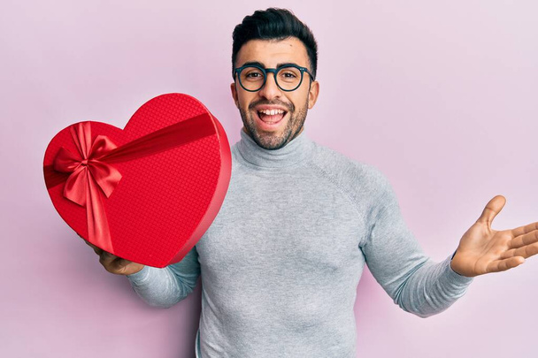 Junger hispanischer Mann mit Valentinstagsgeschenk feiert Errungenschaft mit glücklichem Lächeln und Siegerausdruck mit erhobener Hand  - Foto, Bild