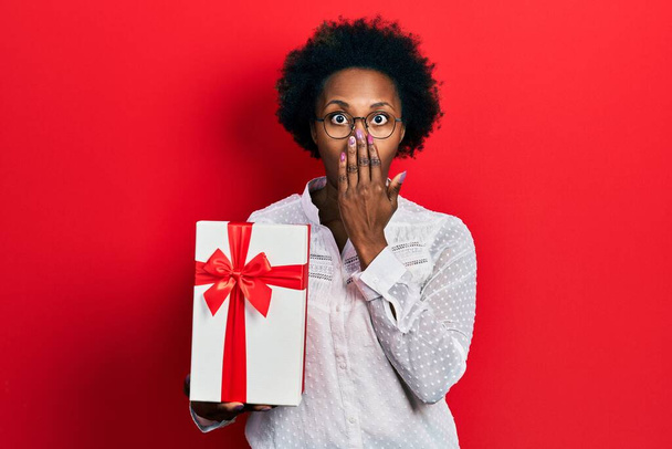 Young african american woman holding gift covering mouth with hand, shocked and afraid for mistake. surprised expression  - Fotó, kép