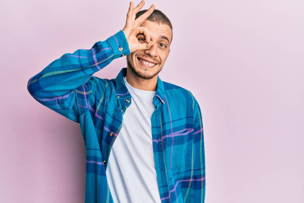 Latino nuori mies yllään rento vaatteita hymyillen onnellinen tekee ok merkki käsin silmään katselee sormien läpi  - Valokuva, kuva