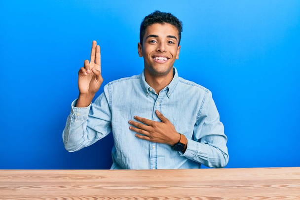 Mladý pohledný africký Američan v neformálním oblečení sedí na stole a usmívá se s rukama na prsou a prsty nad hlavou a přísahá věrnost.  - Fotografie, Obrázek