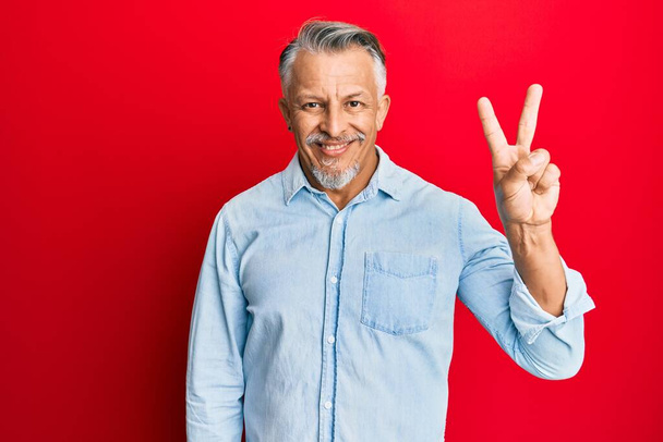 Чоловік середнього віку у сірому одязі, що з'являється і вказує пальцями на номер два, посміхаючись впевнено і щасливо
.  - Фото, зображення