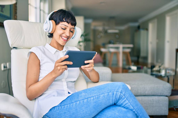 Beautiful brunette woman with short hair at home wearing headphones using touchpad device - Foto, immagini