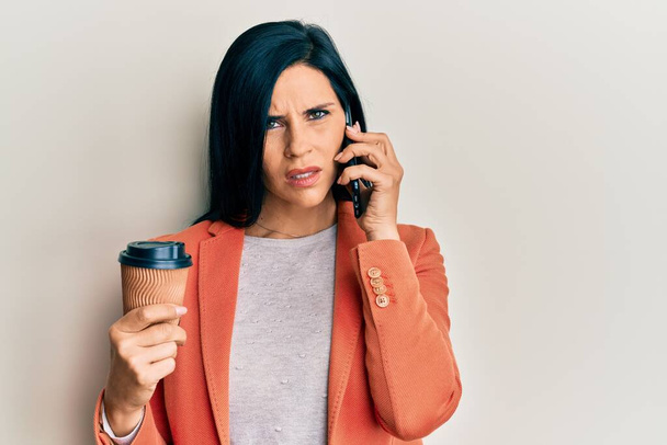 Νεαρή καυκάσια γυναίκα που κάνει συζήτηση μιλώντας στο smartphone κρατώντας τον καφέ στο πρόσωπο σοκ, δείχνοντας σκεπτική και σαρκαστική, έκπληκτη με το ανοιχτό στόμα  - Φωτογραφία, εικόνα