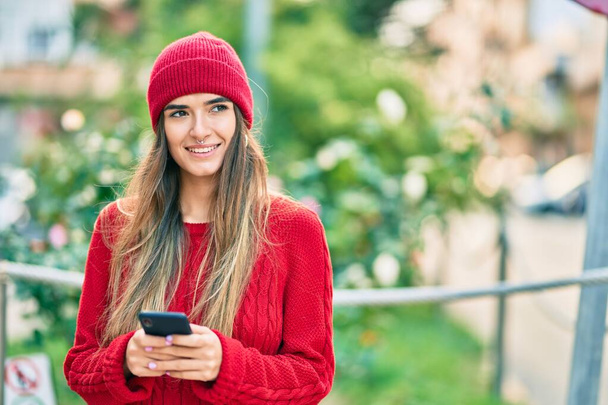 Νεαρή Ισπανίδα γυναίκα που φοράει χειμερινό στυλ χρησιμοποιώντας smartphone στην πόλη. - Φωτογραφία, εικόνα