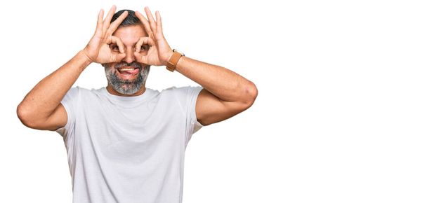 Middle age handsome man wearing casual white tshirt doing ok gesture like binoculars sticking tongue out, eyes looking through fingers. crazy expression.  - Zdjęcie, obraz