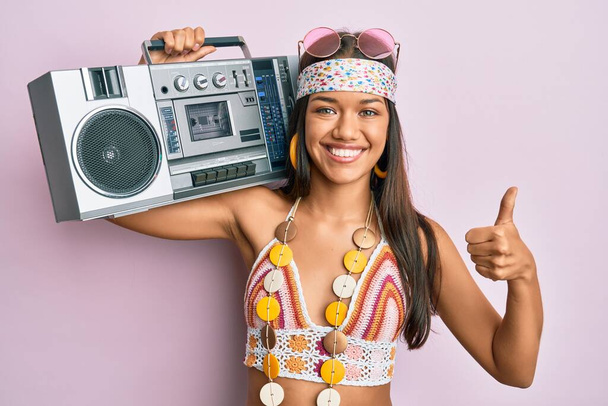Beautiful hispanic woman wearing hippie style holding boom box smiling happy and positive, thumb up doing excellent and approval sign  - Photo, Image