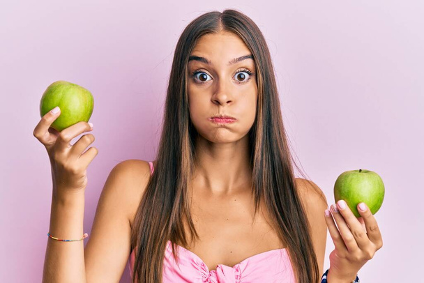 Młoda latynoska kobieta trzymająca zielone jabłka nadyma policzki zabawną twarzą. usta napompowane powietrzem, łapie powietrze.  - Zdjęcie, obraz