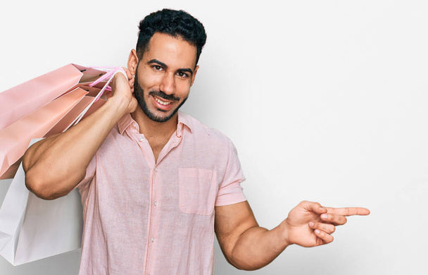 Latino mies parta tilalla ostoskassit hymyillen onnellinen osoittaa kädellä ja sormella sivulle  - Valokuva, kuva