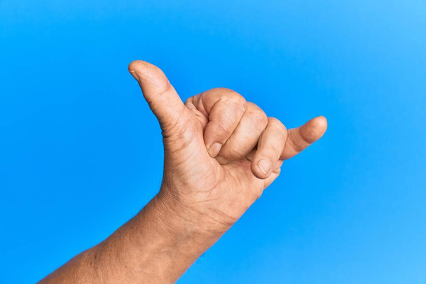 Ręka starszego Hiszpana nad niebieskim, odizolowanym tłem, gestem powitania hawajskiego shaka, symbolem telefonu i komunikacji  - Zdjęcie, obraz