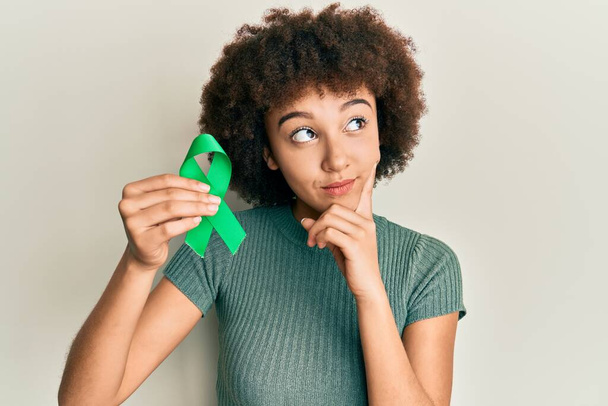 Młoda Latynoska dziewczyna trzyma wsparcie zielona wstążka poważna twarz myśląc o pytaniu z ręki na podbródek, myśląc o mylące pomysł  - Zdjęcie, obraz
