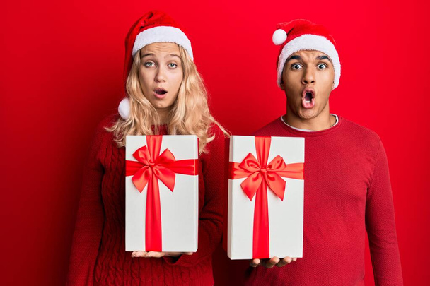 Νεαρό διαφυλετικό ζευγάρι φορώντας χριστουγεννιάτικο καπέλο και κρατώντας ένα δώρο φοβάται και έκπληκτος με ανοιχτό το στόμα για έκπληξη, δυσπιστία πρόσωπο  - Φωτογραφία, εικόνα