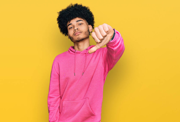 Jonge Afro-Amerikaanse man met afrohaar in casual roze sweater die er ongelukkig en boos uitziet en afwijzing toont en negatief met duimen naar beneden gebaar. slechte uitdrukking.  - Foto, afbeelding