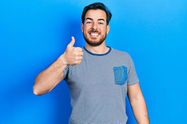 Handsome caucasian man with beard wearing casual striped t shirt doing happy thumbs up gesture with hand. approving expression looking at the camera showing success.  - Fotó, kép