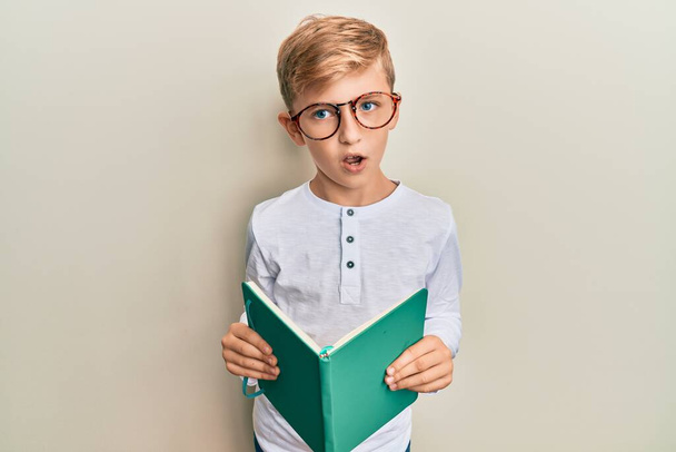 Kis kaukázusi fiú egy könyvet olvas szemüveget visel sokkos arccal, szkeptikusan és szarkasztikusan néz ki, meglepve nyitott szájjal  - Fotó, kép
