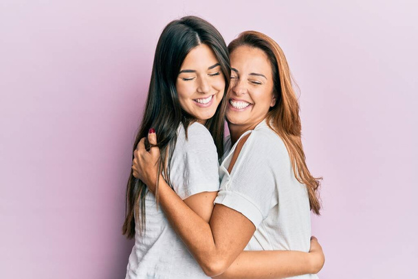 mooie latino moeder en dochter glimlachen gelukkig knuffelen over geïsoleerde roze achtergrond. - Foto, afbeelding