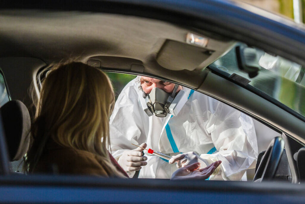 Woman sitting in car, waiting for medical worker to perform drive-thru COVID-19 test, taking nasal swab sample through car window, PCR diagnostic for Coronavirus, doctor in PPE holding test kit. - Fotografie, Obrázek