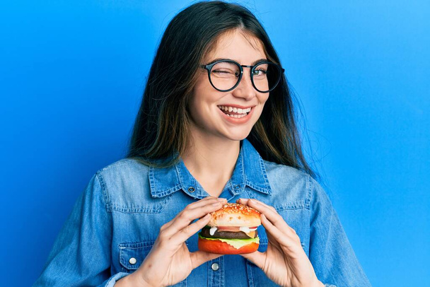 若いですcaucasian女性食べるおいしいです古典的なハンバーガーウィンク見ますザ · カメラとともにセクシー表現,陽気で幸せな顔.  - 写真・画像