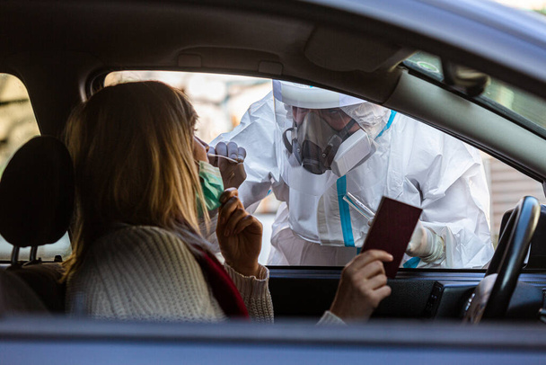 Pracownik medyczny wykonujący test drive-thru COVID-19, pobierający wymaz z nosa od pacjentki przez okno samochodu, diagnostykę PCR, lekarz w apteczce PPE. Trzyma paszporty.. - Zdjęcie, obraz