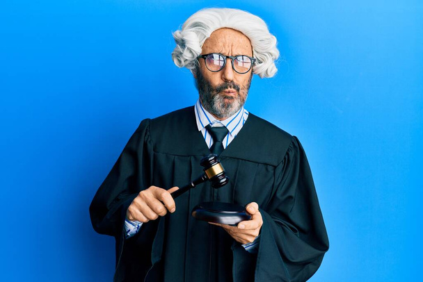 Middle age hispanic man using gavel making fish face with mouth and squinting eyes, crazy and comical.  - Photo, Image