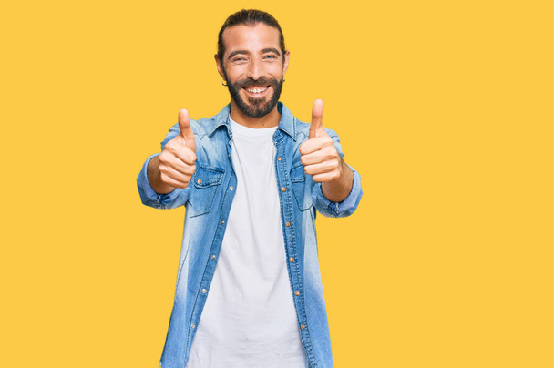 Attractive man with long hair and beard wearing casual denim jacket success sign doing positive gesture with hand, thumbs up smiling and happy. cheerful expression and winner gesture.  - Photo, Image
