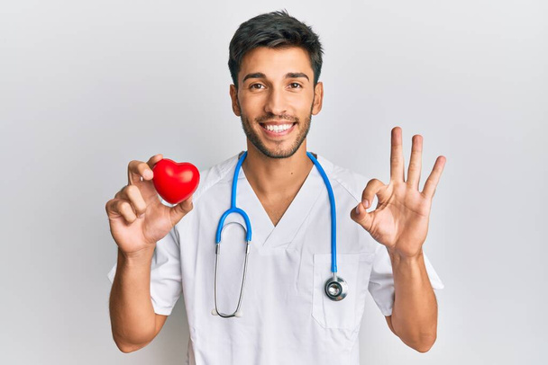 Young handsome man wearing doctor uniform holding heart doing ok sign with fingers, smiling friendly gesturing excellent symbol  - Foto, Imagem