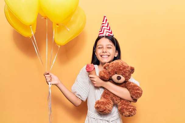 Urocza latynoska dziewczynka w urodzinowym kapeluszu uśmiechnięta. Stojąc z uśmiechem na twarzy trzymając balony i pluszowego misia jedząc lody na odizolowanym żółtym tle - Zdjęcie, obraz