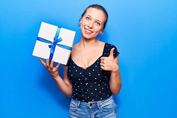 Young blonde woman holding gift smiling happy and positive, thumb up doing excellent and approval sign  - Photo, Image