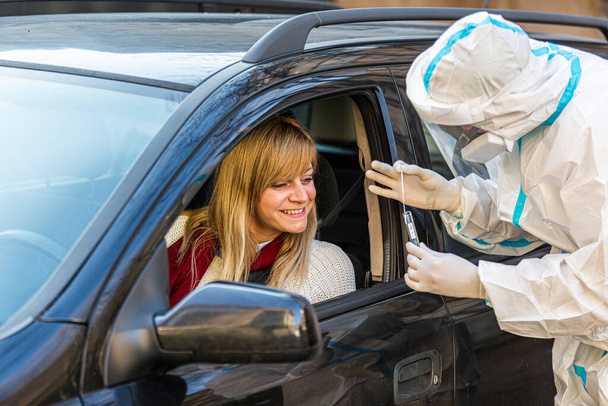 Žena sedí v autě, čeká na zdravotníka, aby provedl test COVID-19, odebírá nosní stěr oknem auta, PCR diagnostika pro Coronavirus, lékař v PPE drží testovací soupravu. - Fotografie, Obrázek