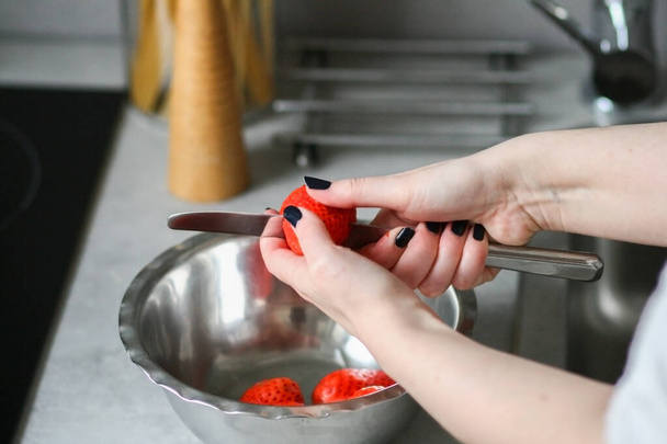 slicing strawberries into a metal bowl, kitchen with an imitation concrete worktop, a woman with painted nails cuts strawberries with a knife, healthy, organic breakfast morning - Fotografie, Obrázek