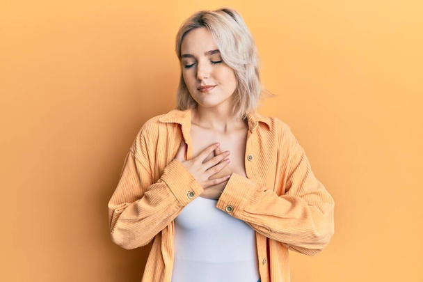 Young blonde girl wearing casual clothes smiling with hands on chest with closed eyes and grateful gesture on face. health concept.  - Photo, Image